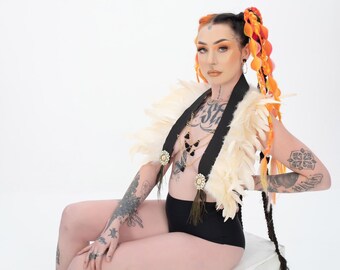 May include: A person with orange and pink hair wearing a white feather boa and a black choker with a gold chain and black pendants. They are also wearing black underwear and have tattoos on their arms and chest.