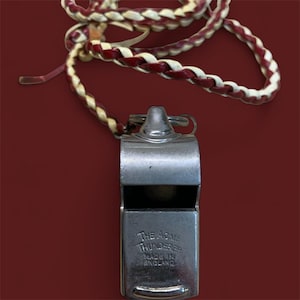 May include: A silver-colored Acme Thunderer whistle with a braided red and white lanyard. The whistle has the text "THE ACME THUNDERER MADE IN ENGLAND" engraved on the front. The background is a solid maroon color.