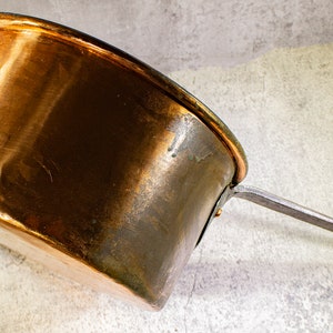 May include: A copper saucepan with a long, straight handle. The pot has a rounded shape with a slightly flared rim. The copper has a warm, reddish-brown color with some darker areas, suggesting age or use. The handle is made of a lighter-colored metal.