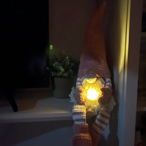 Puede incluir: Un gnomo de crochet rosa con un patr&oacute;n de rayas blancas y rosas en las piernas. El gnomo sostiene una peque&ntilde;a luz en sus manos, que ilumina su rostro.