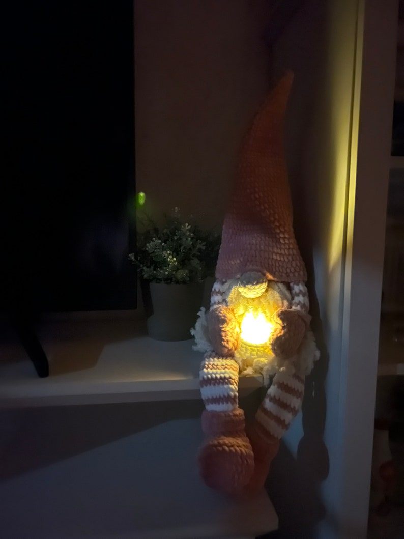 Puede incluir: Un gnomo de crochet rosa con un patr&oacute;n de rayas blancas y rosas en las piernas. El gnomo sostiene una peque&ntilde;a luz en sus manos, que ilumina su rostro.