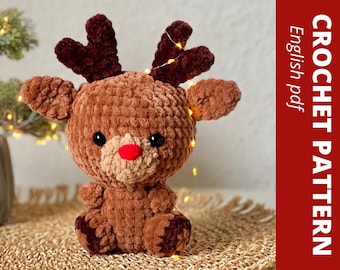 May include: A soft, brown crocheted reindeer toy with dark brown antlers and a red nose. The reindeer has black eyes and is sitting on a woven mat. The image includes the text "CROCHET PATTERN English pdf".