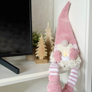 Puede incluir: Un gnomo de crochet rosa con rayas blancas en las piernas y un gorro de crochet blanco con un pomp&oacute;n blanco en la parte superior. El gnomo est&aacute; sentado en un estante blanco.