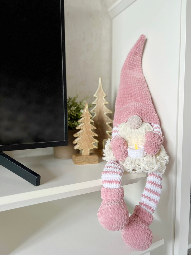 Puede incluir: Un gnomo de crochet rosa con rayas blancas en las piernas y un gorro de crochet blanco con un pomp&oacute;n blanco en la parte superior. El gnomo est&aacute; sentado en un estante blanco.