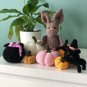 K&ouml;nnte beinhalten: Eine Sammlung von geh&auml;kelten Halloween-Dekorationen, darunter eine braune Fledermaus, eine schwarze Spinne, ein Kessel und vier K&uuml;rbisse in verschiedenen Farben.