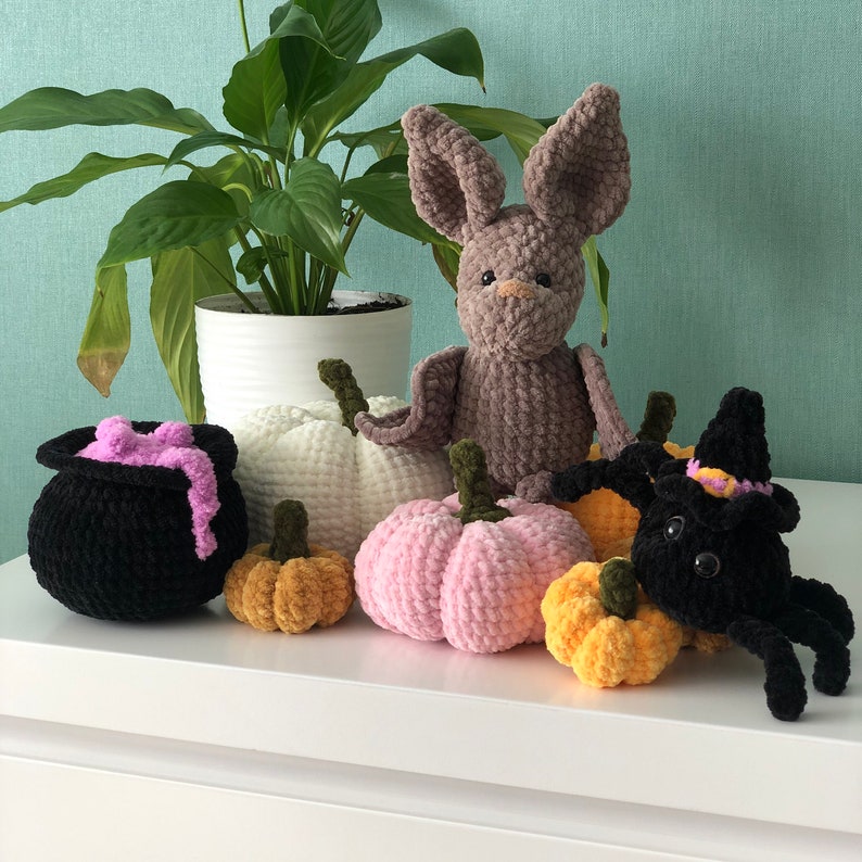K&ouml;nnte beinhalten: Eine Sammlung von geh&auml;kelten Halloween-Dekorationen, darunter eine braune Fledermaus, eine schwarze Spinne, ein Kessel und vier K&uuml;rbisse in verschiedenen Farben.