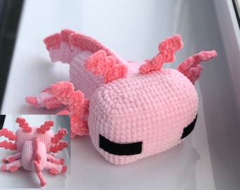 May include: Pink crocheted axolotl plush toy with black eyes. The axolotl is lying on its side with its head turned to the right.