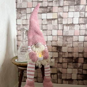 Puede incluir: Un gnomo de crochet rosa con barba blanca sosteniendo una vela peque&ntilde;a. El gnomo est&aacute; sentado en una mesa de madera con una botella de vidrio decorativa en el fondo.