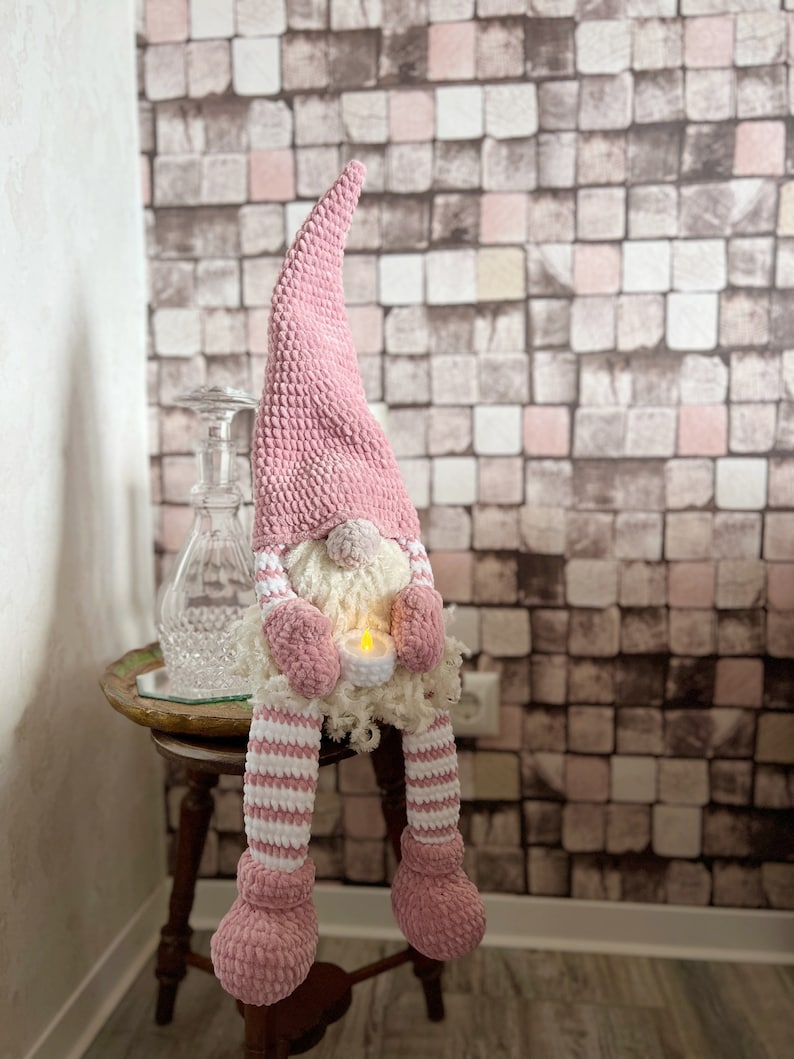 Puede incluir: Un gnomo de crochet rosa con barba blanca sosteniendo una vela peque&ntilde;a. El gnomo est&aacute; sentado en una mesa de madera con una botella de vidrio decorativa en el fondo.