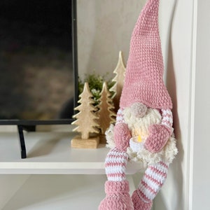 Puede incluir: Un gnomo de crochet rosa con barba blanca y piernas a rayas blancas y rosas est&aacute; sentado en un estante blanco. El gnomo sostiene una vela peque&ntilde;a en sus manos. Hay dos peque&ntilde;os &aacute;rboles de madera detr&aacute;s del gnomo.