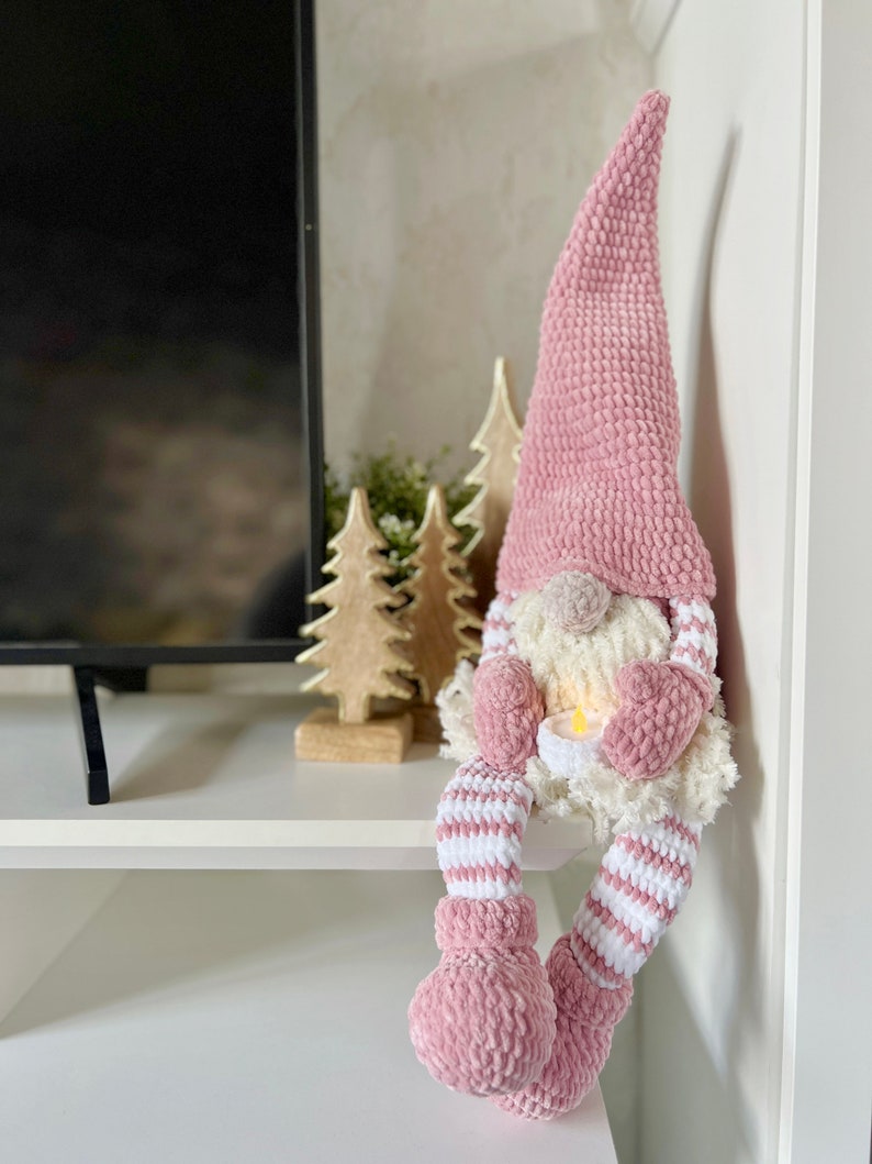 Puede incluir: Un gnomo de crochet rosa con barba blanca y piernas a rayas blancas y rosas est&aacute; sentado en un estante blanco. El gnomo sostiene una vela peque&ntilde;a en sus manos. Hay dos peque&ntilde;os &aacute;rboles de madera detr&aacute;s del gnomo.