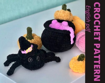 May include: A black crocheted spider wearing a witch's hat with a pink and yellow band. The spider is surrounded by crocheted pumpkins in orange and pink.