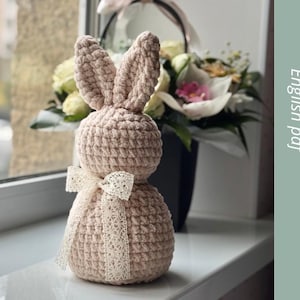 May include: A crocheted pink bunny rabbit with a white lace bow. The bunny is sitting on a windowsill with a bouquet of flowers in the background. Crochet pattern.