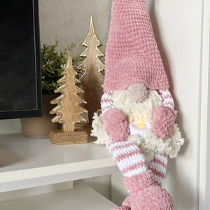 Puede incluir: Un gnomo de crochet rosa con un gorro de pomp&oacute;n blanco y piernas a rayas blancas y rosas. El gnomo est&aacute; sentado en un estante blanco con dos &aacute;rboles de Navidad de madera detr&aacute;s.