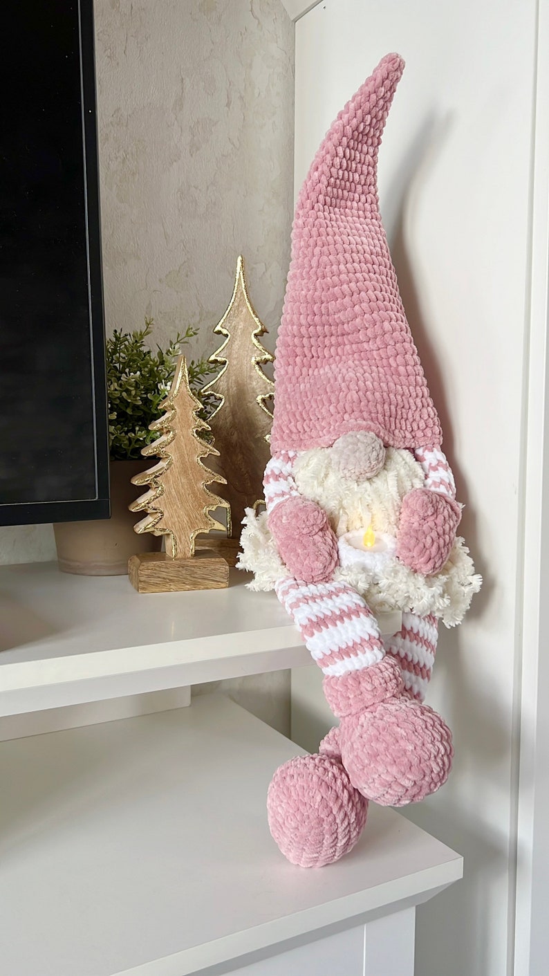 Puede incluir: Un gnomo de crochet rosa con un gorro de pomp&oacute;n blanco y piernas a rayas blancas y rosas. El gnomo est&aacute; sentado en un estante blanco con dos &aacute;rboles de Navidad de madera detr&aacute;s.