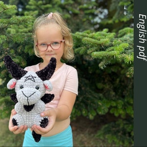 May include: A child holds a gray and black crocheted goat plush toy. The goat has a pentagram on its forehead and black horns. The child is wearing a pink headband and glasses. The background is a green bush.