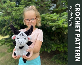 May include: A child holds a gray and black crocheted goat plush toy. The goat has a pentagram on its forehead and black horns. The child is wearing a pink headband and glasses. The background is a green bush.
