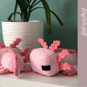 Peut inclure: Trois peluches axolotl en crochet rose avec des détails rose et blanc. Les peluches sont posées sur une surface blanche. Patron de crochet en anglais pdf.
