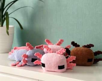May include: Three crocheted axolotl plushies in pink, purple, and brown. The plushies have pink fins and black eyes.