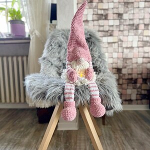 Puede incluir: Un gnomo de crochet rosa con un cuerpo a rayas blanco y rosa se sienta en una silla de pelo gris. El gnomo tiene una barba blanca y un gorro rosa.