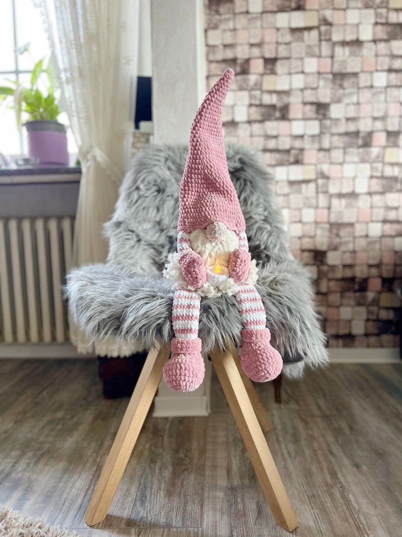 Puede incluir: Un gnomo de crochet rosa con un cuerpo a rayas blanco y rosa se sienta en una silla de pelo gris. El gnomo tiene una barba blanca y un gorro rosa.