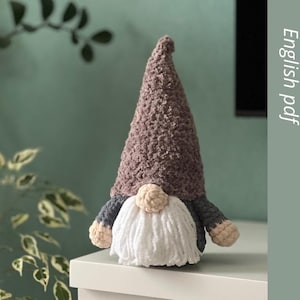May include: Crochet pattern for a gnome with a brown hat, white beard, and grey and beige accents. The pattern is available as a digital download in English PDF format.