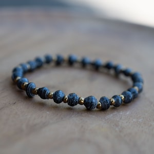 May include: A black and gold paper bead bracelet. The beads are made of rolled paper and have a textured look. The bracelet is strung on a gold cord.