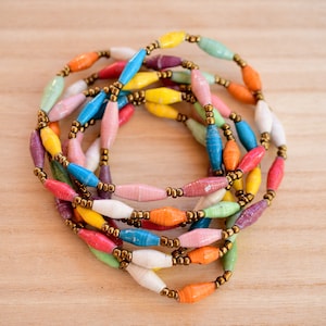 May include: A colorful necklace made of paper beads in various hues, including yellow, orange, pink, blue, and green. The beads are strung together with gold-colored metal spacers, creating a vibrant and eye-catching accessory.