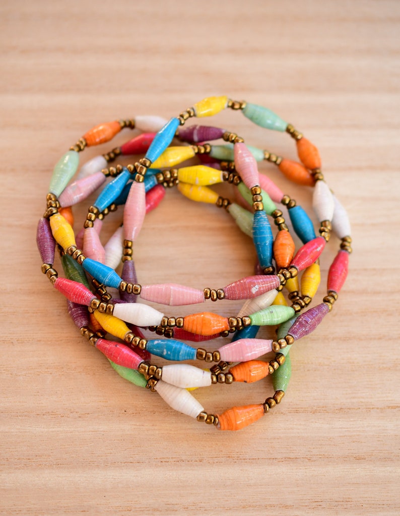May include: A colorful necklace made of paper beads in various hues, including yellow, orange, pink, blue, and green. The beads are strung together with gold-colored metal spacers, creating a vibrant and eye-catching accessory.