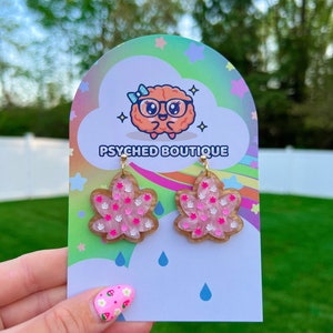 May include: Pink and white flower-shaped earrings with gold-colored hooks. The earrings are displayed on a card with the text "PSYCHED BOUTIQUE" and a cartoon brain logo. The background features a colorful design with stars and rain drops.