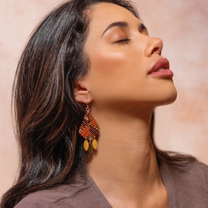 Puede incluir: Primer plano de una mujer con pendientes estampados geométricos. Los pendientes son principalmente naranjas, rojos y negros, con detalles dorados. La mujer lleva una blusa marrón.