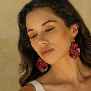 May include: A woman wearing a gold necklace and matching earrings. The earrings and pendant are leaf-shaped with a red, black, and yellow plaid pattern. The woman is wearing a white top.