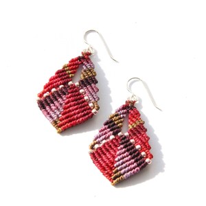 May include: A pair of red, purple and gold diamond-shaped earrings with silver hooks. The earrings are made using a macrame technique and have small white beads.