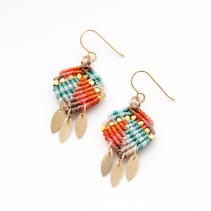 May include: A pair of macrame earrings with a geometric design. The earrings are made with teal, orange, and pink threads, and have gold-coloured beads. Gold-coloured leaf charms hang from the bottom, and gold-coloured ear wires complete the design.