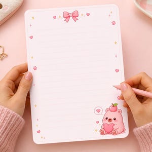 May include: A pink notepad with a bow and heart design, featuring a cute bear illustration holding a heart. The notepad has lined pages and is being written on with a pink pen. The overall theme is stationery and cute designs.