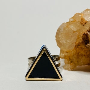 May include: A silver-toned ring featuring a large, black triangle with a gold-colored border. The geometric design is set against a white background, with a natural, light-brown rock formation in the background. The ring has a double-band design.