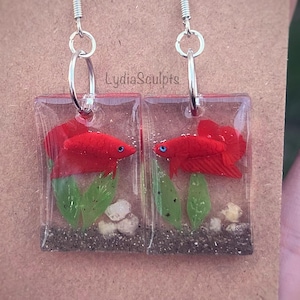 May include: A pair of silver earrings with red betta fish in clear resin. The fish are surrounded by green plants and white gravel. The earrings are rectangular in shape.