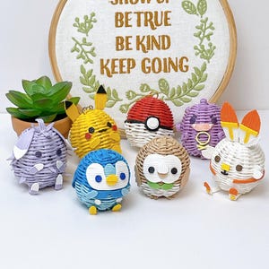 May include: A collection of colorful, woven character figurines, including Pikachu, a Poké Ball, and others. The figurines are displayed in front of an embroidered hoop with the text "SHOW UP BE TRUE BE KIND KEEP GOING". A small potted succulent is also present.