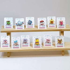 May include: A display of colorful, pixelated character pins on cards, arranged on a two-tiered wooden shelf. Each pin features a unique design, with various colors and shapes. The cards have the text "PAPER TIMES".