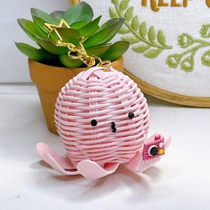 May include: A pink woven octopus keychain with a gold star-shaped clasp. The octopus has black eyes and a small camera. The keychain is next to a small potted succulent. The background includes a framed embroidery hoop.