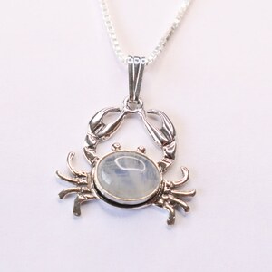 May include: A silver crab pendant necklace with a moonstone cabochon. The crab is detailed with claws and legs. The moonstone is iridescent and has a milky white color.