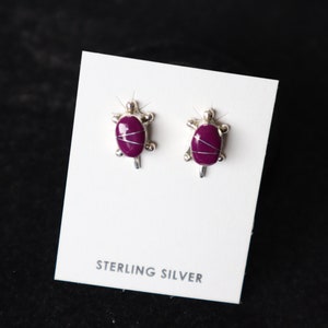 May include: A pair of sterling silver earrings featuring a purple turtle design. The earrings are displayed on a white card with the text "STERLING SILVER".