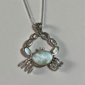 May include: A silver-toned necklace featuring a crab pendant. The pendant has a pearlescent shell body and is surrounded by oval-shaped accents. The crab's claws are detailed, and the necklace hangs on a delicate chain, creating a nautical theme.