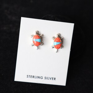 May include: A pair of sterling silver earrings featuring a turtle design with coral and turquoise inlay. The earrings are displayed on a white card with the text "STERLING SILVER".