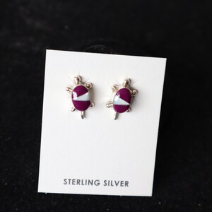 May include: A pair of sterling silver earrings featuring a purple and white turtle design. The earrings are displayed on a white card with the text "STERLING SILVER".
