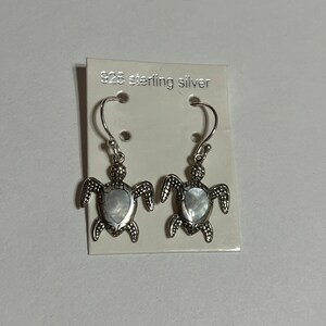 May include: A pair of silver turtle earrings with a mother of pearl inlay. The earrings are on a white card with the text "925 sterling silver".