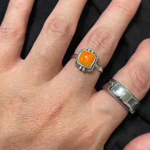 May include: A silver ring with an orange square stone and a second silver band ring. The orange stone ring has a decorative border. The rings are on a person's hand.