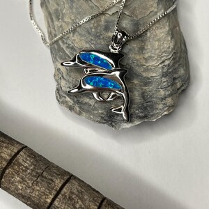 May include: A silver dolphin pendant necklace with blue opal inlays. The pendant features two dolphins, one above the other, on a delicate chain. The necklace is displayed on a grey stone, with a wooden element in the foreground.