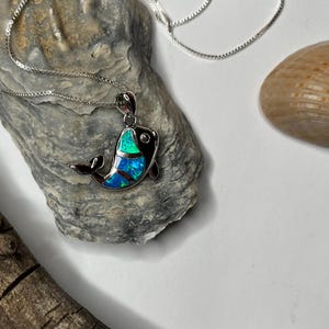 May include: A silver chain necklace with a blue and green opal fish pendant. The pendant is on a rock-like surface.