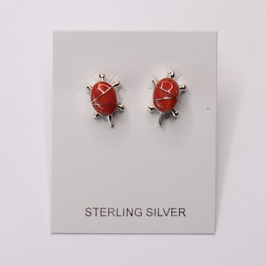 May include: A pair of sterling silver earrings featuring red coral turtle designs. The earrings are displayed on a white card with the text "STERLING SILVER".
